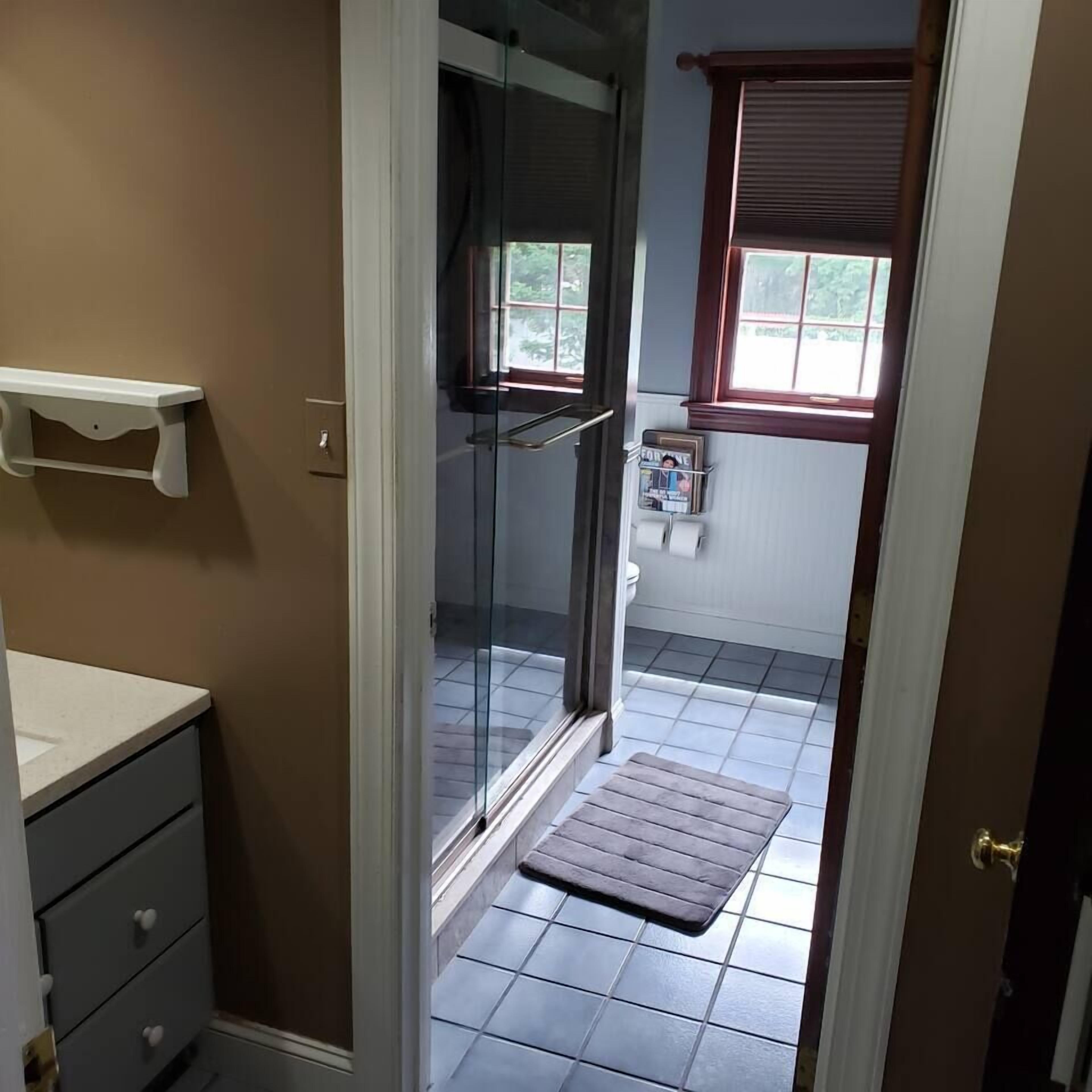 A bathroom with a glass shower enclosure, a small window with blinds, a towel rack, and a white shelf.
