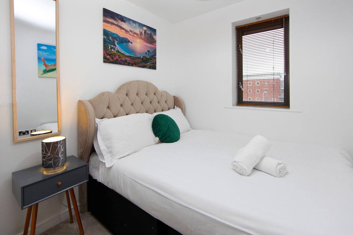 A bedroom with a bed, a bedside table with a lamp, a painting on the wall, and a window with blinds.