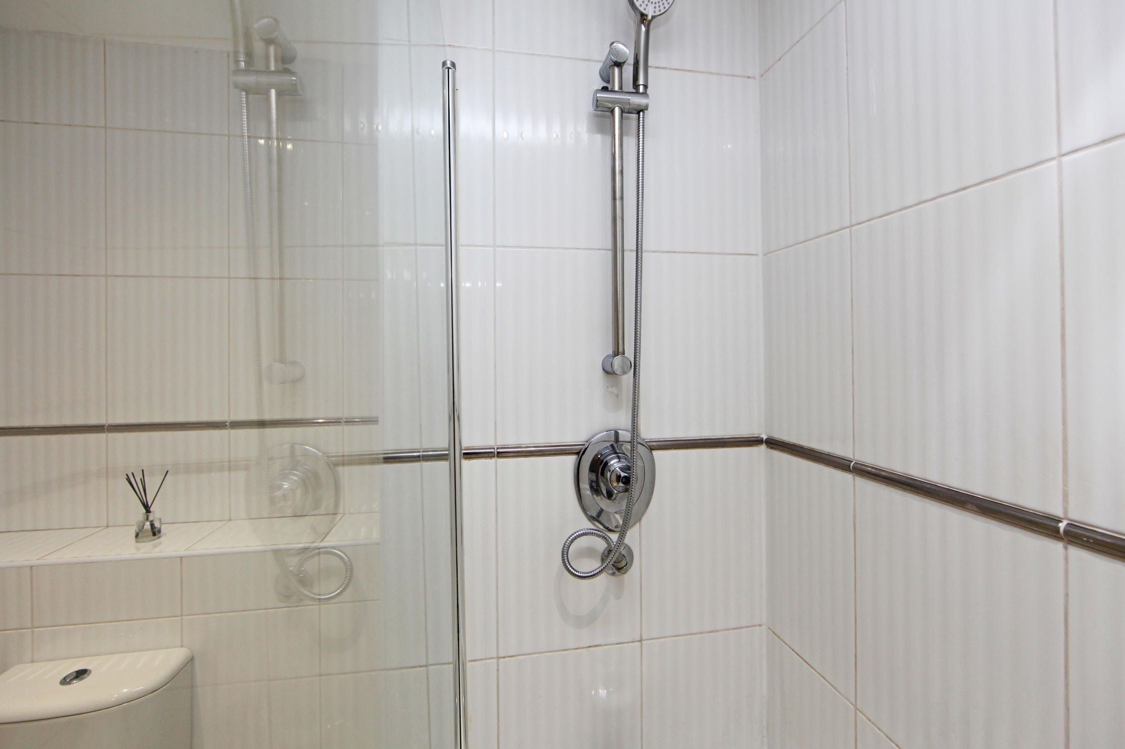 A bathroom with a glass-enclosed shower, a toilet, and a shelf with a glass jar and reed diffuser.