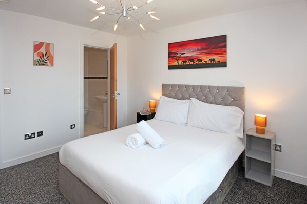 A neatly made bed with white linens and a rolled towel, a tufted headboard, two bedside lamps, and a wall painting of a sunset with silhouettes of elephants.