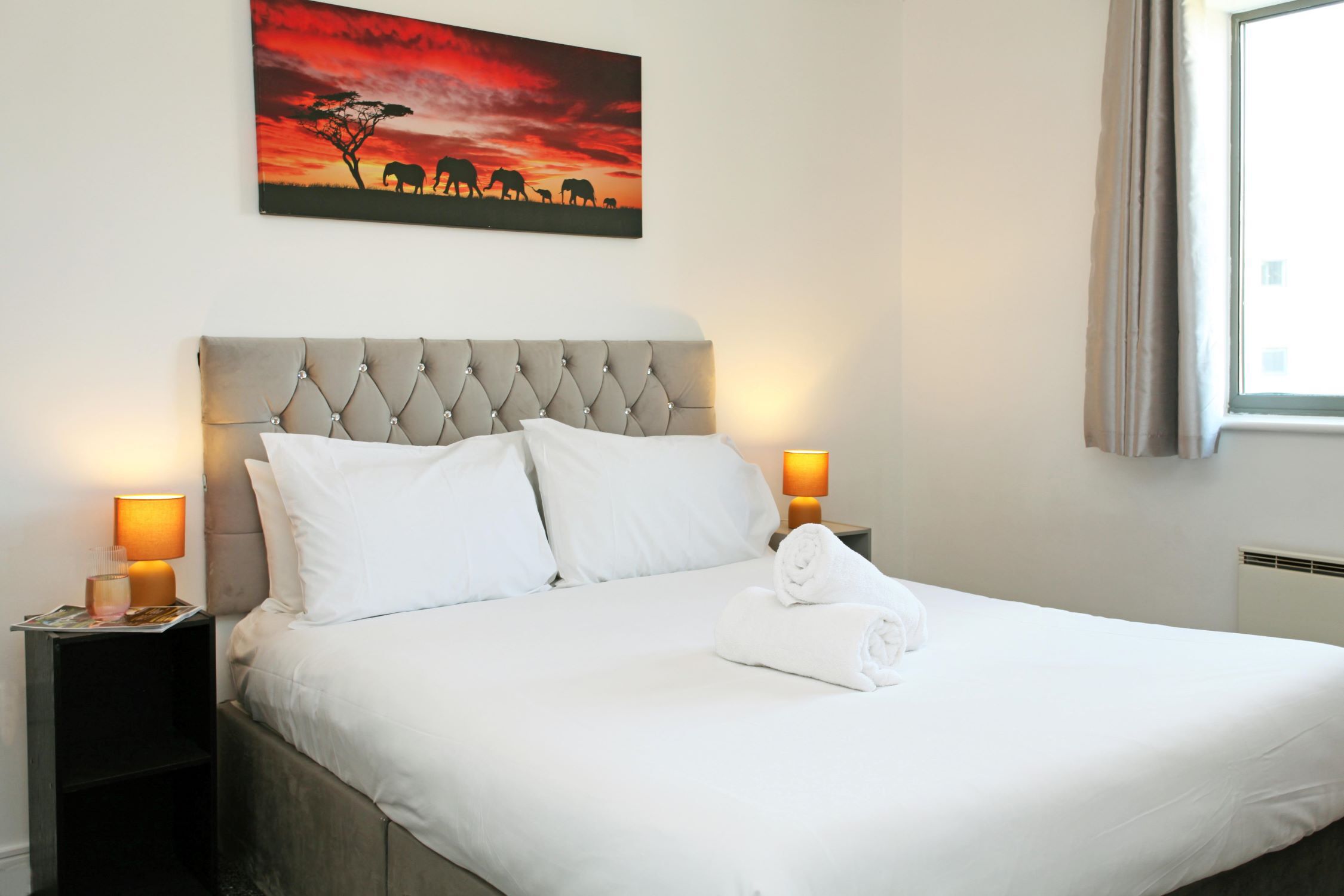 Een netjes opgemaakt bed met witte lakens, twee bedlampjes en een schilderij van olifanten aan de muur.