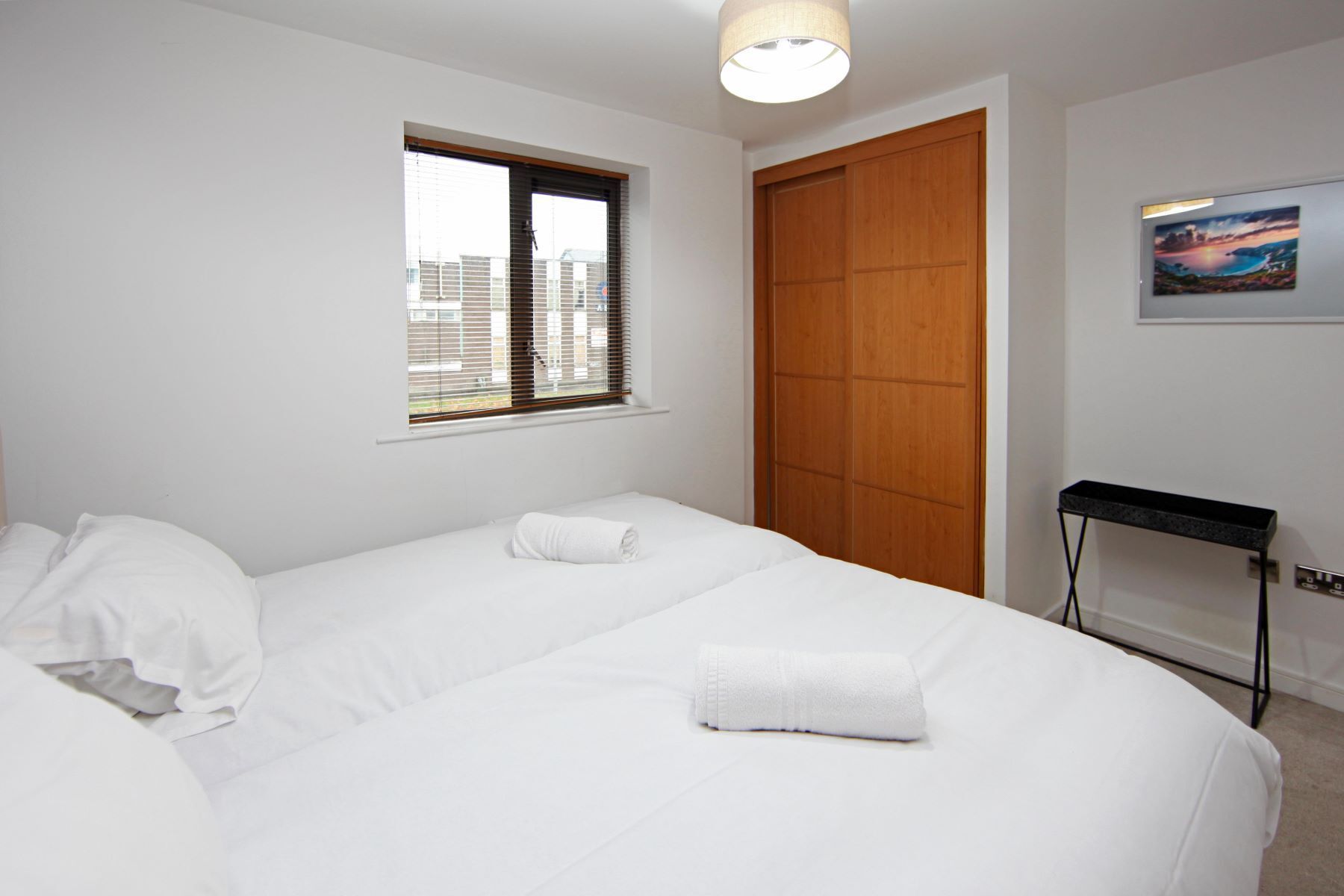 Una habitación de hotel con dos camas, una ventana con persianas, una puerta de madera, un cuadro en la pared y una mesita con lámpara.