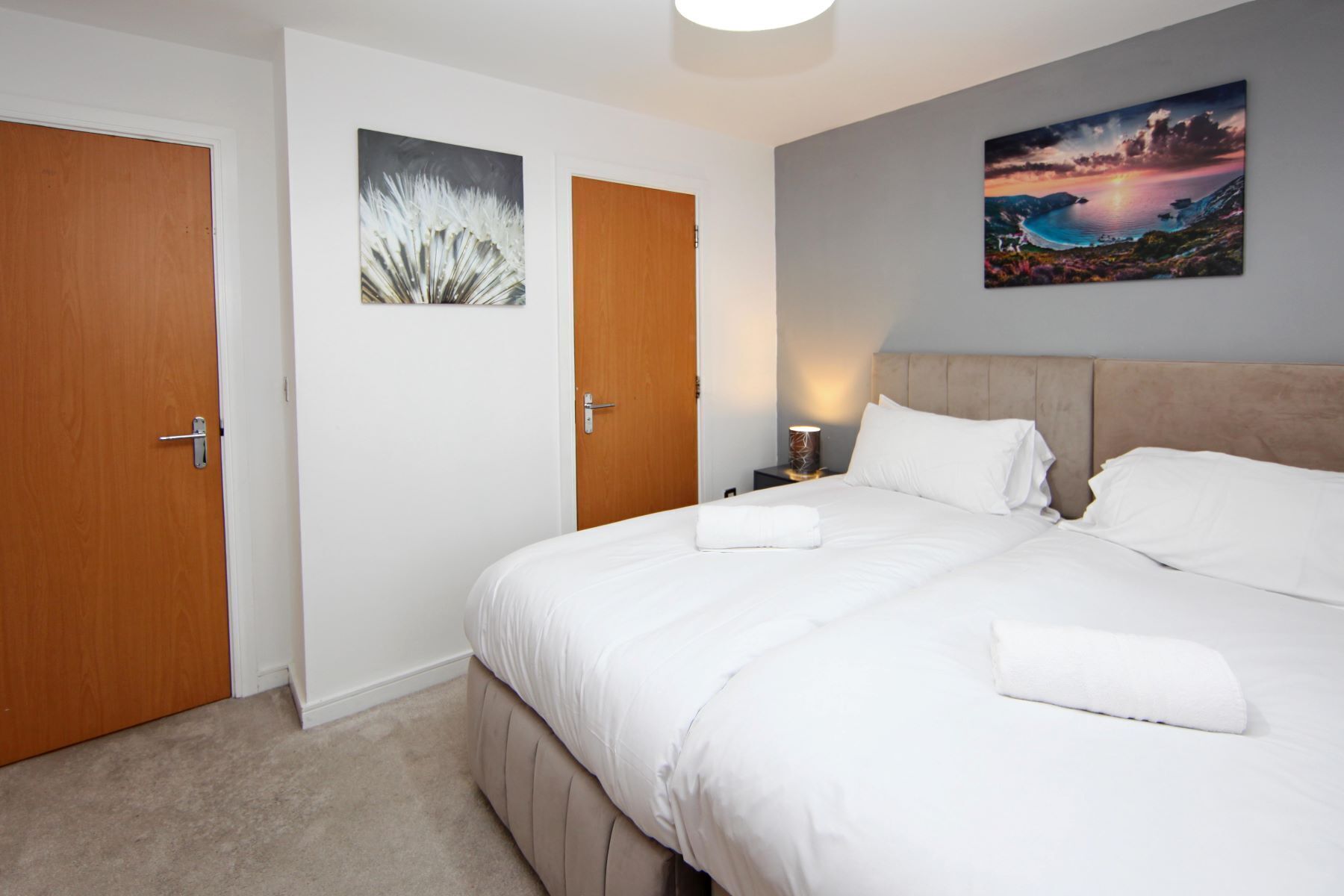 A hotel room with a large bed, two doors, a nightstand with a lamp, and two wall art pieces.