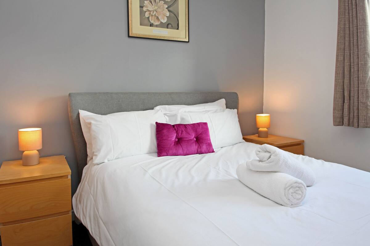 A neatly made bed with white linens and a single purple pillow, flanked by two bedside lamps on wooden nightstands.
