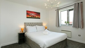A bedroom with a large bed, two bedside tables with lamps, a window with curtains, and a painting on the wall. - Albion Street Apartments (Wolverhampton)