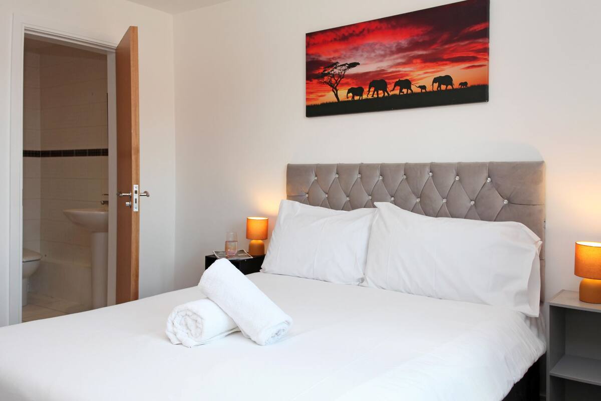 A hotel room with a bed, white linens, a tufted headboard, a painting of elephants, and two bedside lamps.