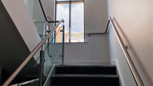A modern staircase with glass railings and a view of the outside through a large window. - Albion Street Apartments (Wolverhampton)