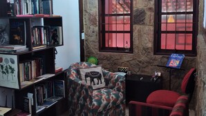 Un coin lecture confortable avec une bibliothèque, un pouf à motifs, un fauteuil rouge et un mur en pierre agrémenté de deux fenêtres.