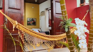 Un hamac est installé devant un bâtiment rouge, sur lequel on peut voir un ventilateur de plafond, une porte et un climatiseur fixé au mur.