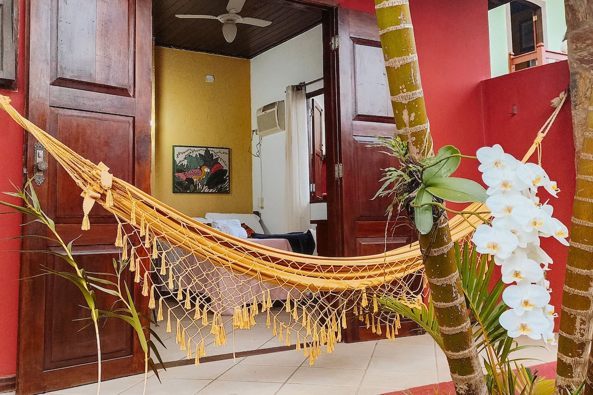 A hammock in front of a red building with a ceiling fan, a door, and a wall-mounted air conditioner.
