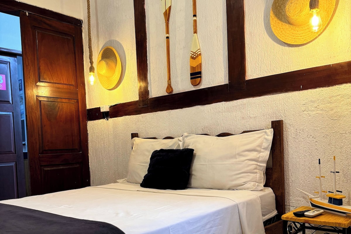 A neatly made bed with white linens and a black throw blanket, a wooden headboard, and a small bedside table with a lamp and a remote control.
