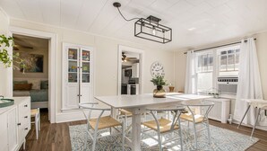 A dining area with a white table and chairs, a patterned rug, and a kitchen in the background. - Lighthouse Cottage: 1 block from the beach, 2 blocks to downtown! (Ludington)