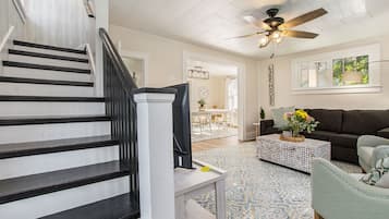 Un escalier avec des rampes noires, un ventilateur de plafond, un salon avec un canapé foncé, une table basse et un tapis à motifs.