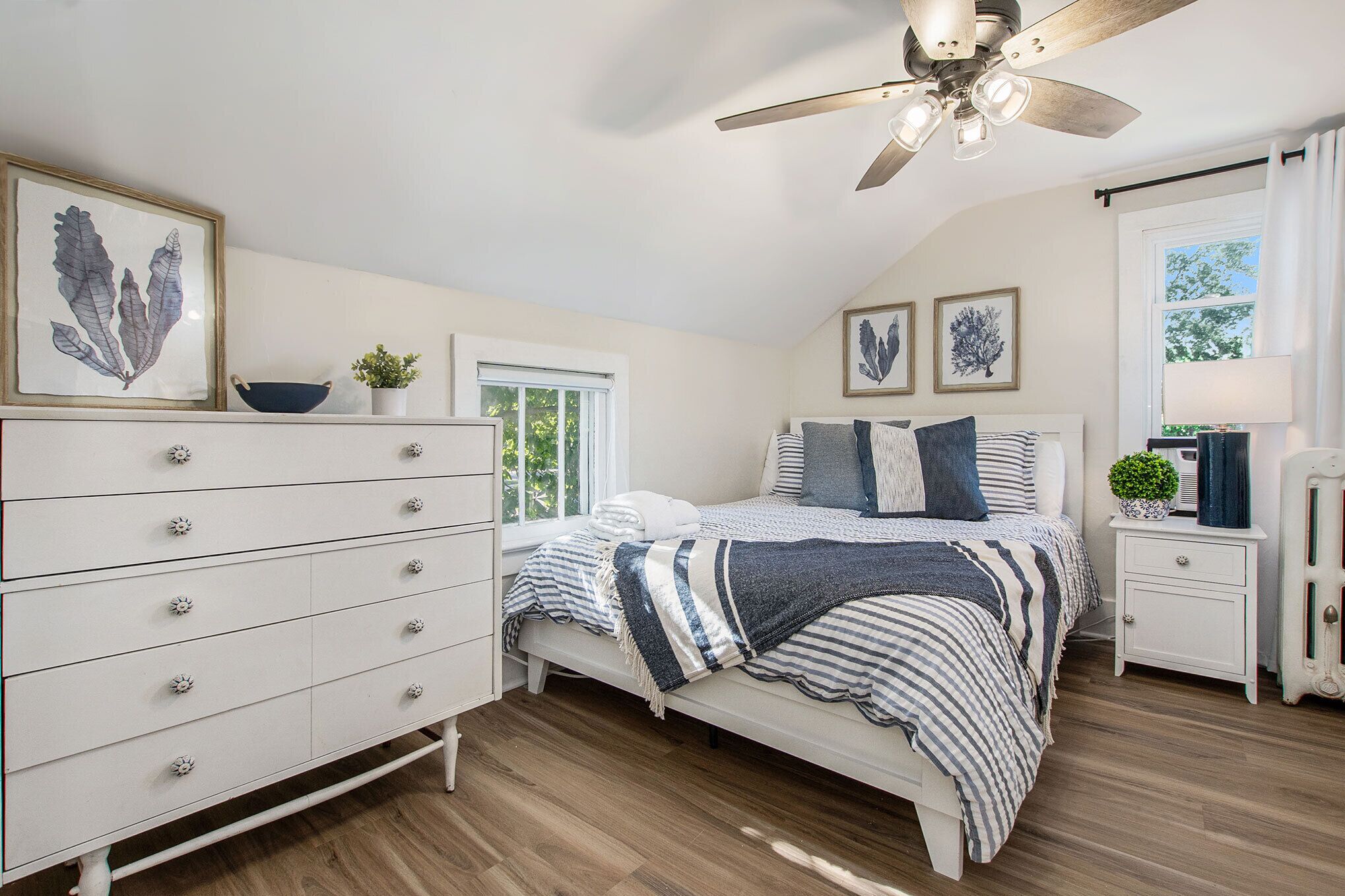 A bedroom with a bed, dresser, nightstand, ceiling fan, and artwork on the walls.