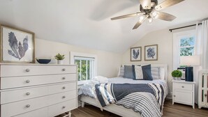 A bedroom with a bed, dresser, nightstand, ceiling fan, and artwork on the walls. - Lighthouse Cottage: 1 block from the beach, 2 blocks to downtown! (Ludington)