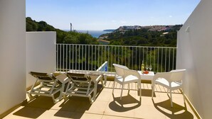 Un balcón con sillas y una mesa blancos, con vistas a la costa, edificios y vegetación.