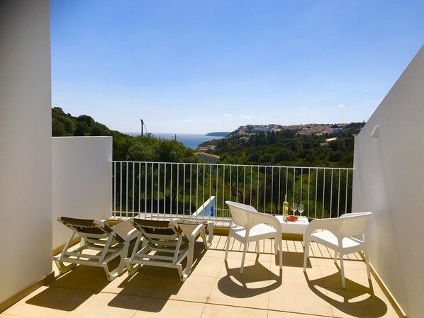 A balcony with white chairs and a table, overlooking a coastal view with buildings and greenery.