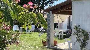 Ein Patio mit weißen Stühlen und einem Tisch unter einer Pergola, umgeben von üppigem Grün und blühenden Pflanzen.