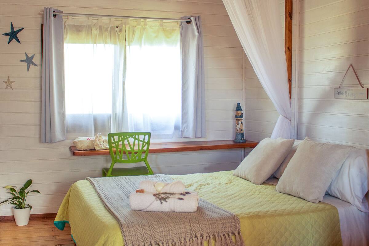 A bedroom with a bed, a window with curtains, a green chair, a potted plant, and a wall-mounted decoration.