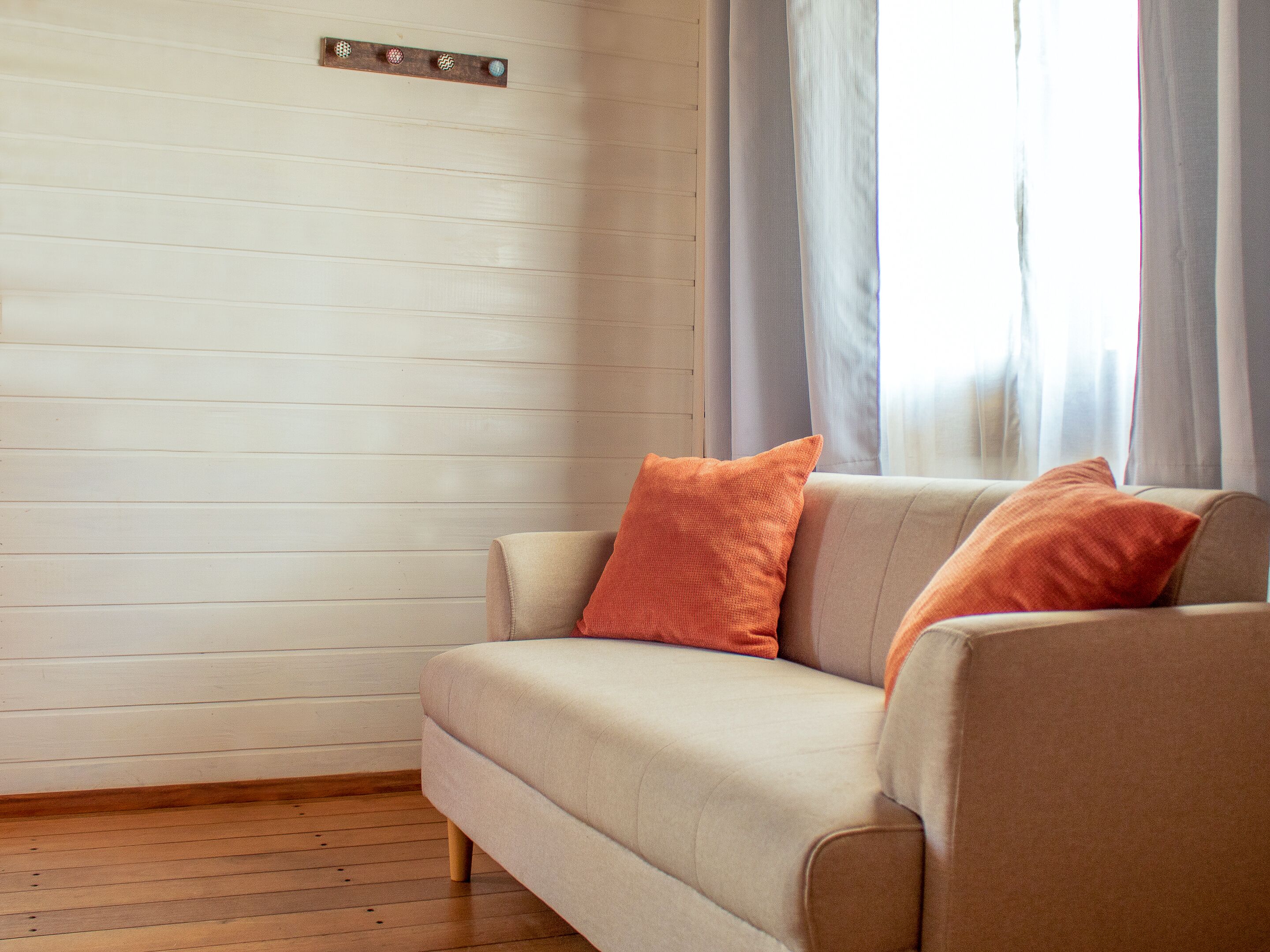 Una sala de estar con un sofá color beige, dos cojines anaranjados, suelo de madera y una pared de tono claro con un panel de madera horizontal.