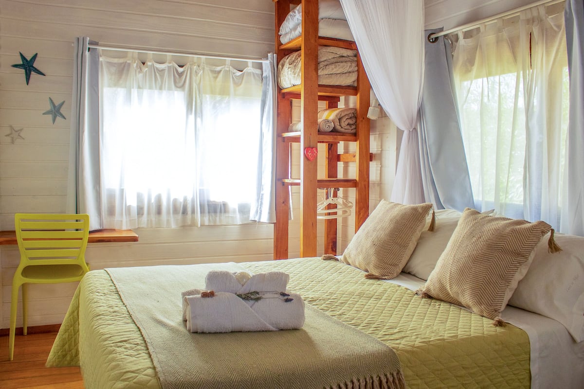 A room with a bed, a wooden shelf with towels, a yellow chair, and a window with sheer curtains.