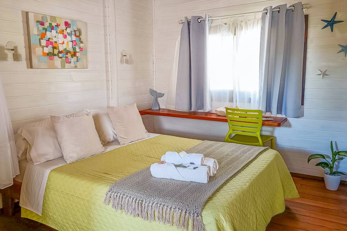 A cozy bedroom with a bed, a window with curtains, a chair, a small table, and a wall decoration.
