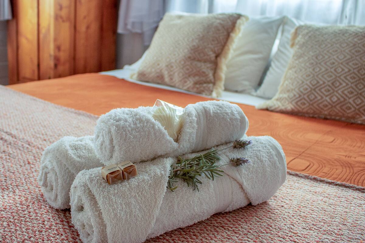 A neatly made bed with white towels and a sprig of rosemary, a wooden headboard, and a patterned throw on a textured orange bedspread.
