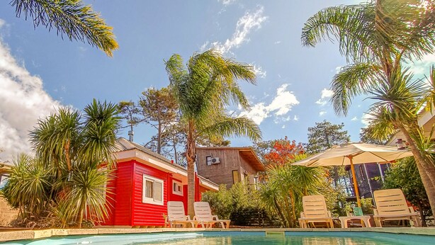 A pool area with lounge chairs, a red building, and palm trees. - Bon Amour Punta del Diablo (Punta del Diablo)
