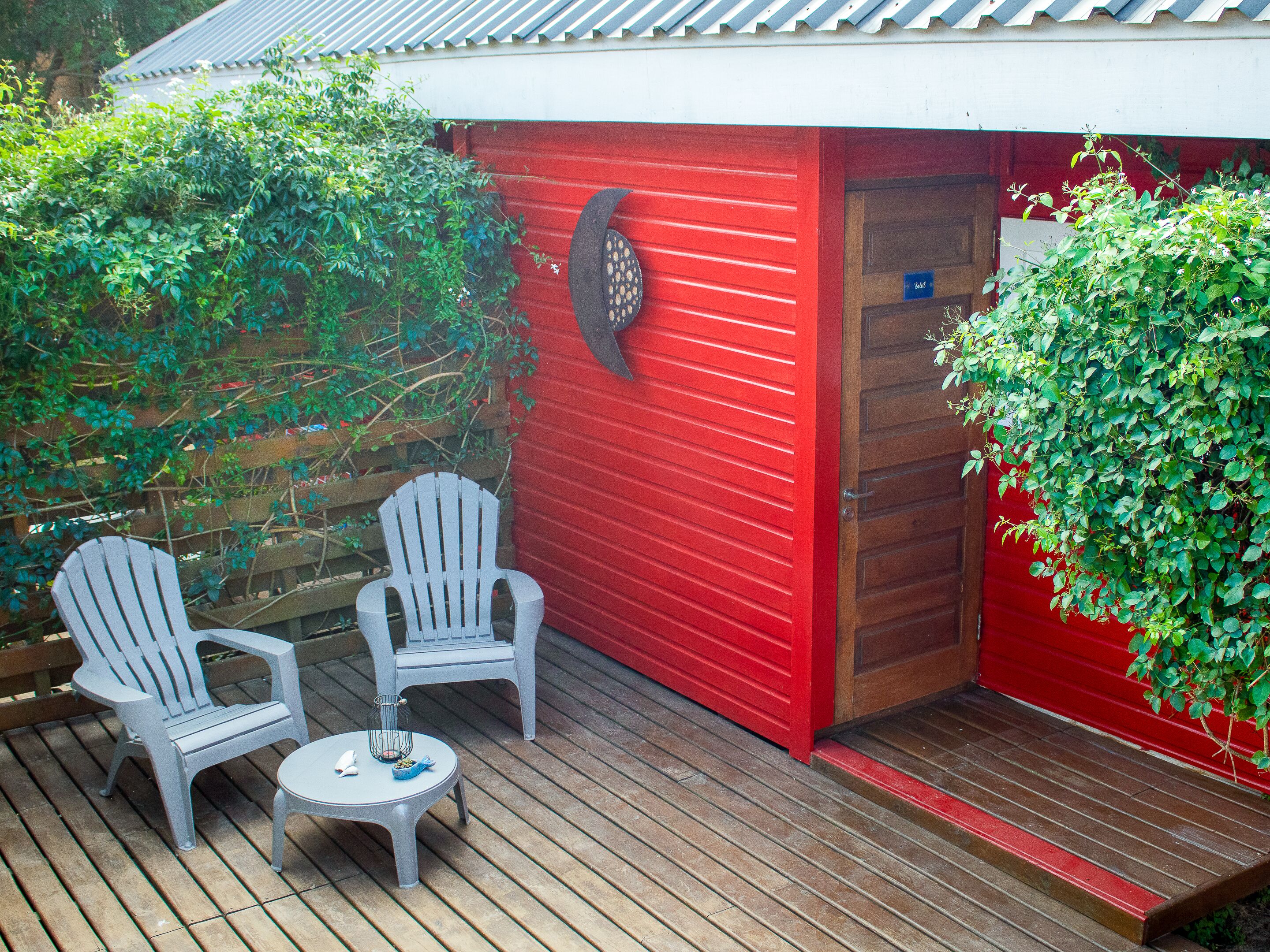 Ein rotes Gebäude mit einer Holztür und einer kleinen Veranda, auf der zwei weiße Stühle und ein runder Tisch stehen.