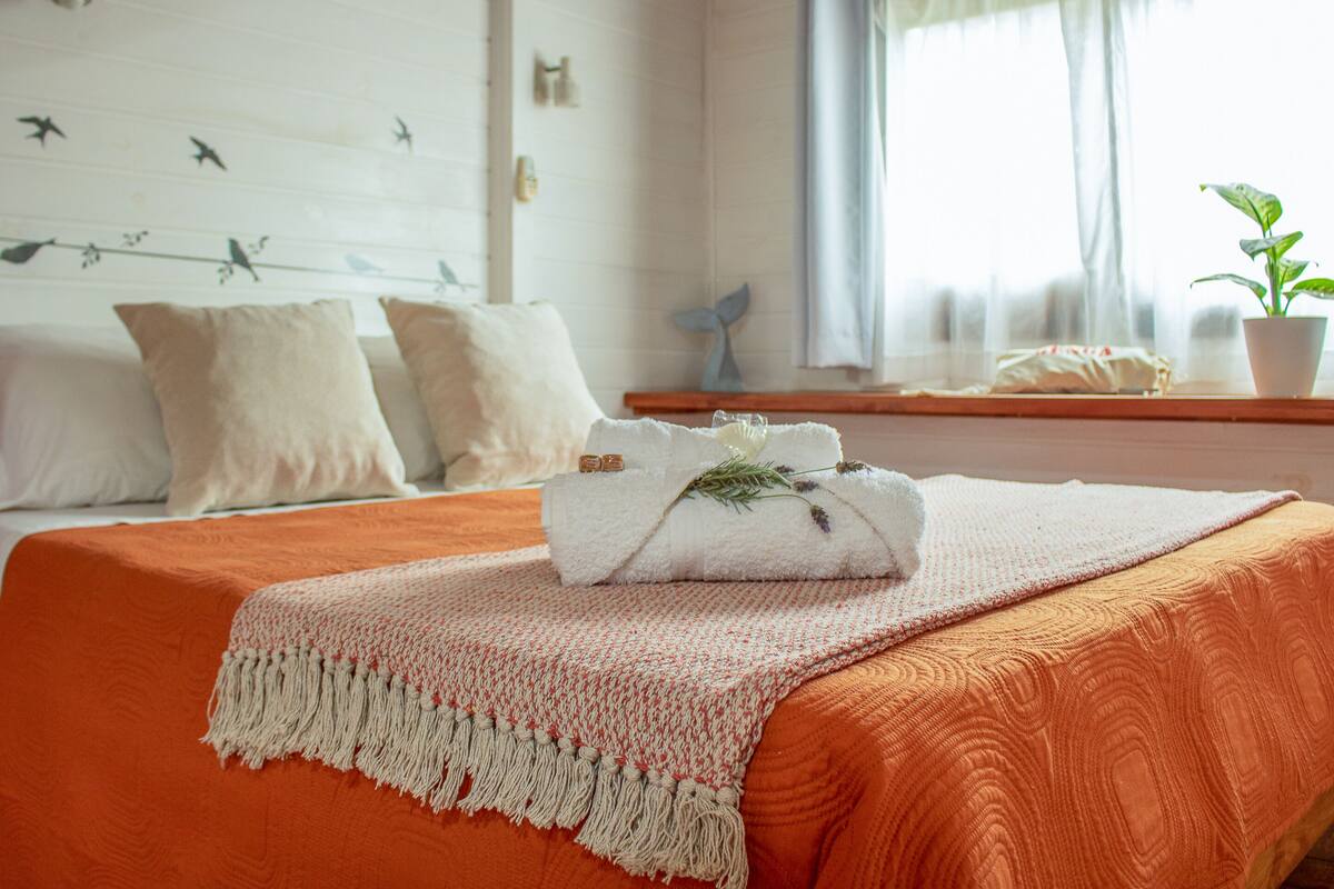 A neatly made bed with an orange bedspread, a white towel with a decorative leaf, and a white wall with bird decorations.