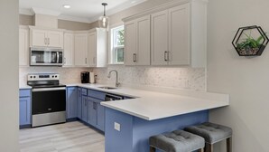A modern kitchen with white cabinets, a stainless steel oven, a microwave, and a dishwasher. There is a large island with stools and a hanging plant. - New! 5 min to beach! Clean, modern and spacious-King mattress on Primary room  (Pensacola)