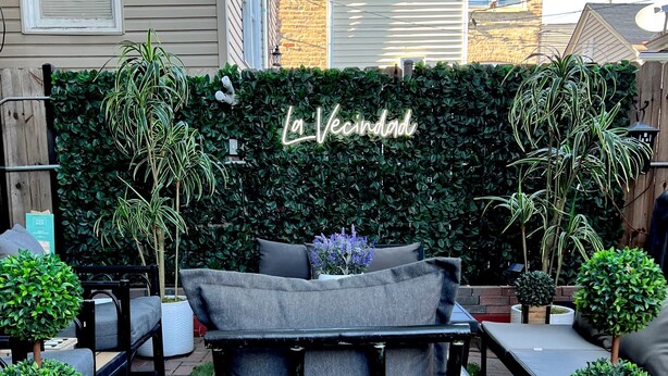 A patio area with a black metal chair, a grey sofa, and a green hedge with a neon sign reading "La Vecindad".