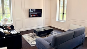 A modern living room with a flat-screen TV mounted on the wall, a dark wood floor, a grey sofa, and a coffee table. - Gîte Vichy - 4 Chambres, Situé Dans le Parc du Château de Charmeil (Charmeil)