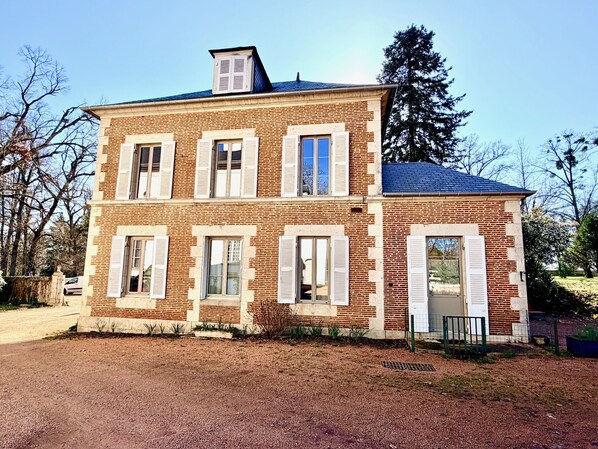 A two-story brick house with white shutters, a small attached garage, and a gravel driveway. - Gîte Vichy - 4 Chambres, Situé Dans le Parc du Château de Charmeil (Charmeil)