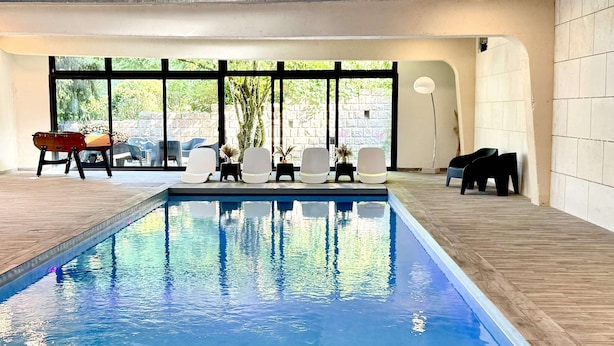 An indoor swimming pool with a wooden deck, lounge chairs, and a view of the outdoors through large windows. - Gîte Vichy - 4 Chambres, Situé Dans le Parc du Château de Charmeil (Charmeil)