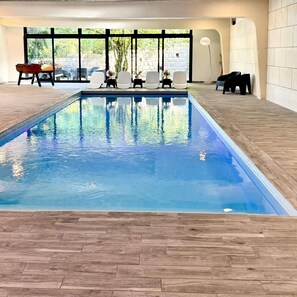 An indoor swimming pool with a wooden deck, lounge chairs, and a view of the outdoors through large windows. - Gîte Vichy - 4 Chambres, Situé Dans le Parc du Château de Charmeil (Charmeil)