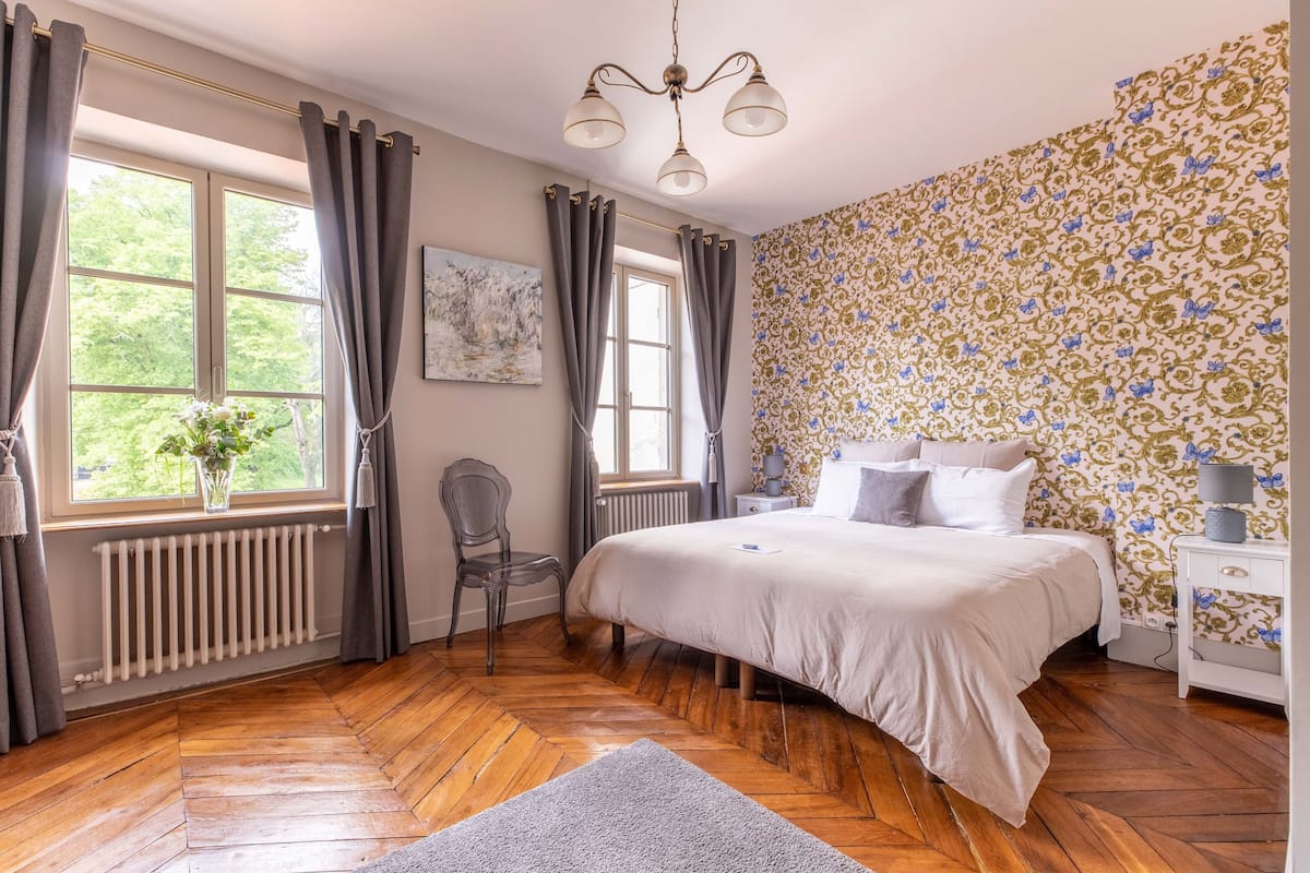 Ein Schlafzimmer mit einem großen Bett, einem Heizkörper, einem Sessel und einem Fenster mit Vorhängen.