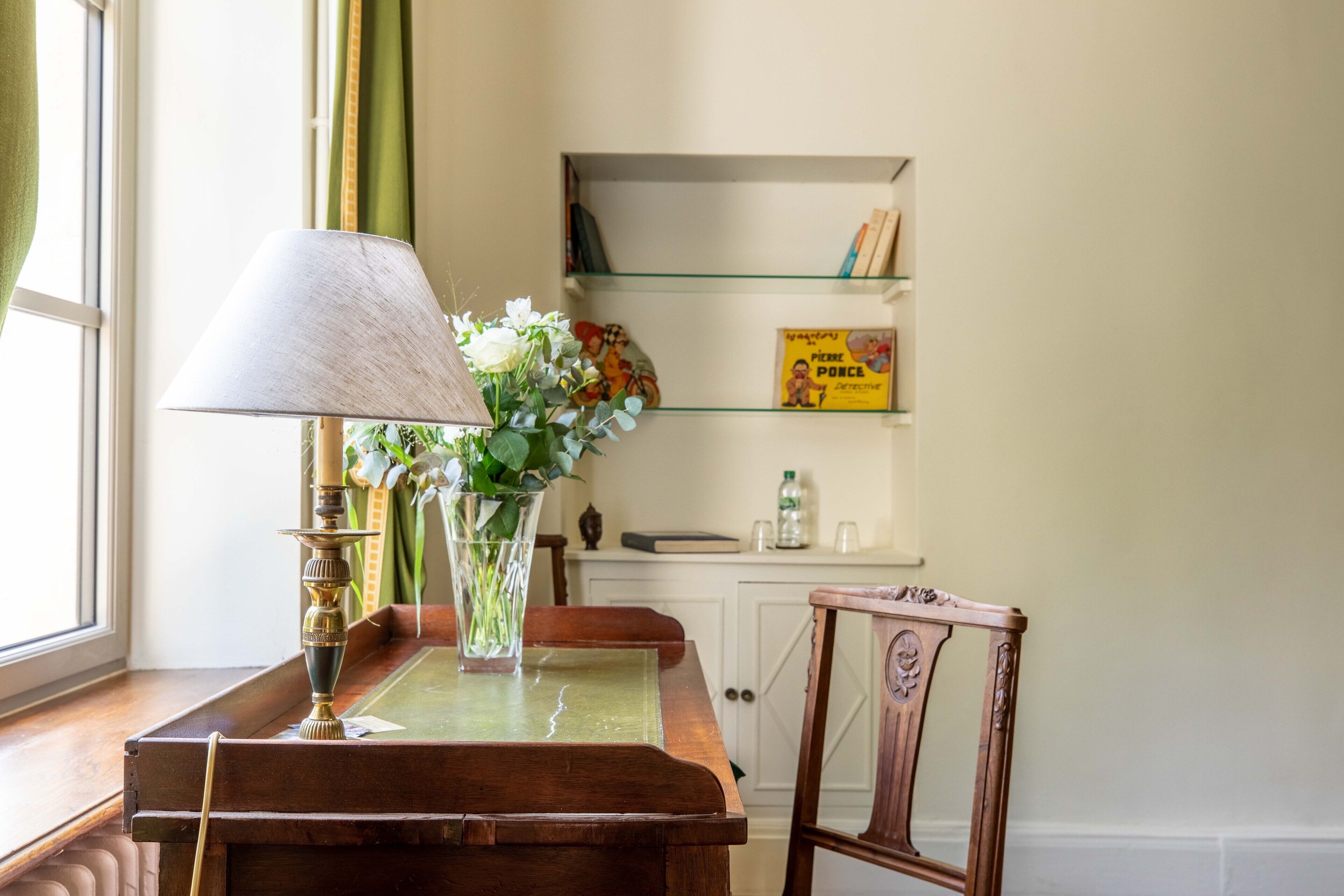 A wooden table with a lamp and a vase of flowers, a chair, and a bookshelf with books and a box.