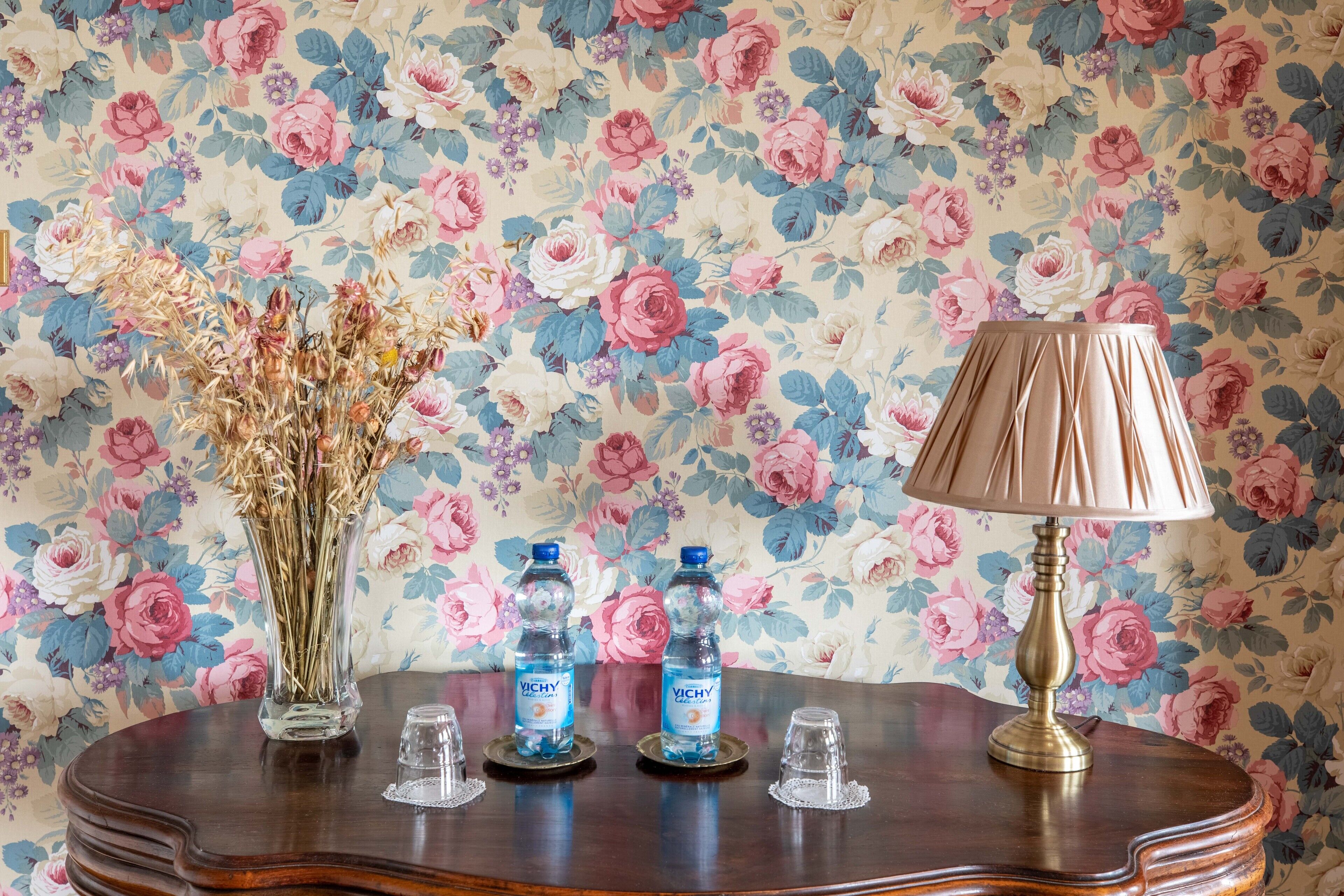 A wooden table with a floral wallpaper background, a vase with dried flowers, two water bottles, and a lamp.