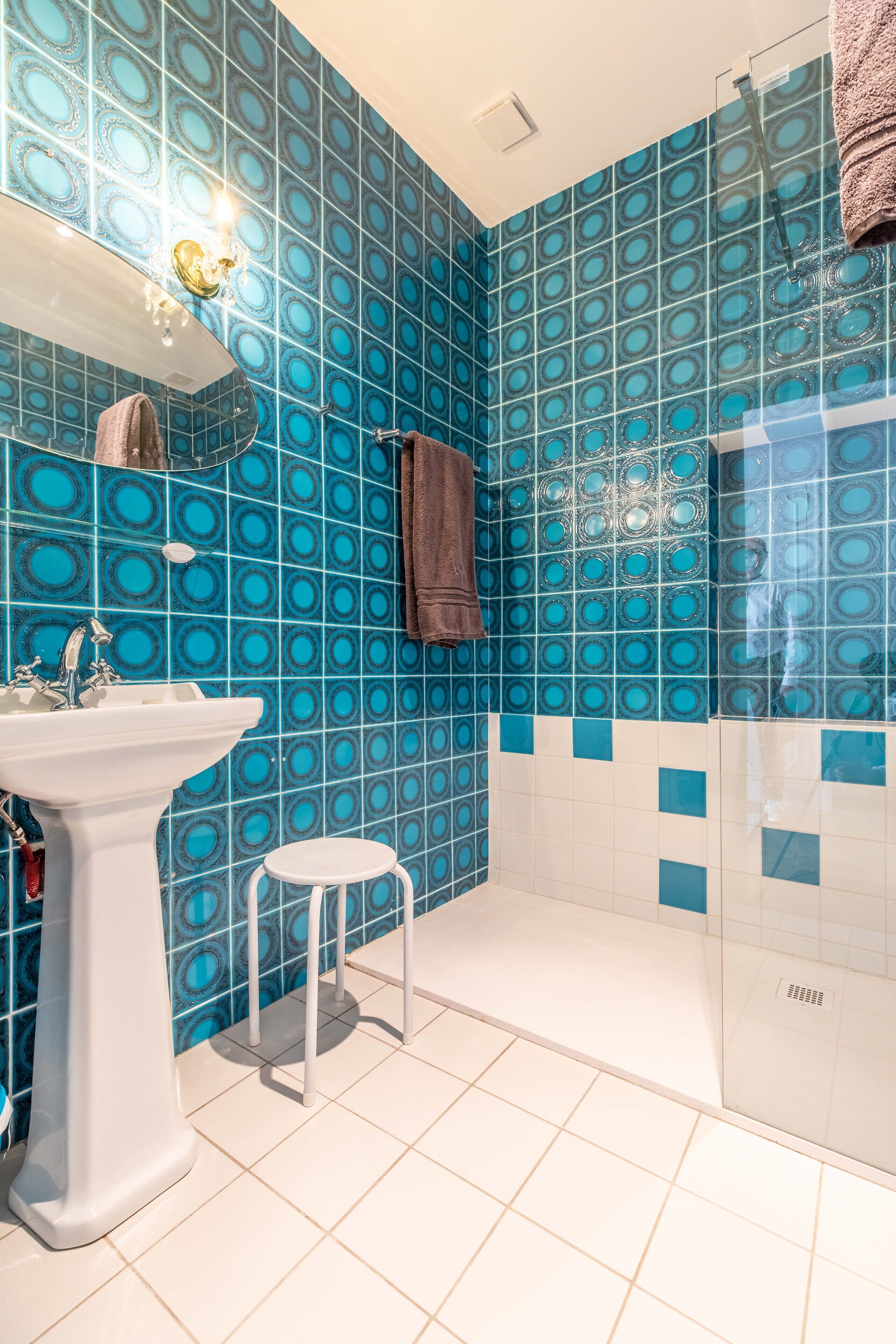 A bathroom with blue patterned tiles, a white pedestal sink, a small stool, and a glass shower enclosure.