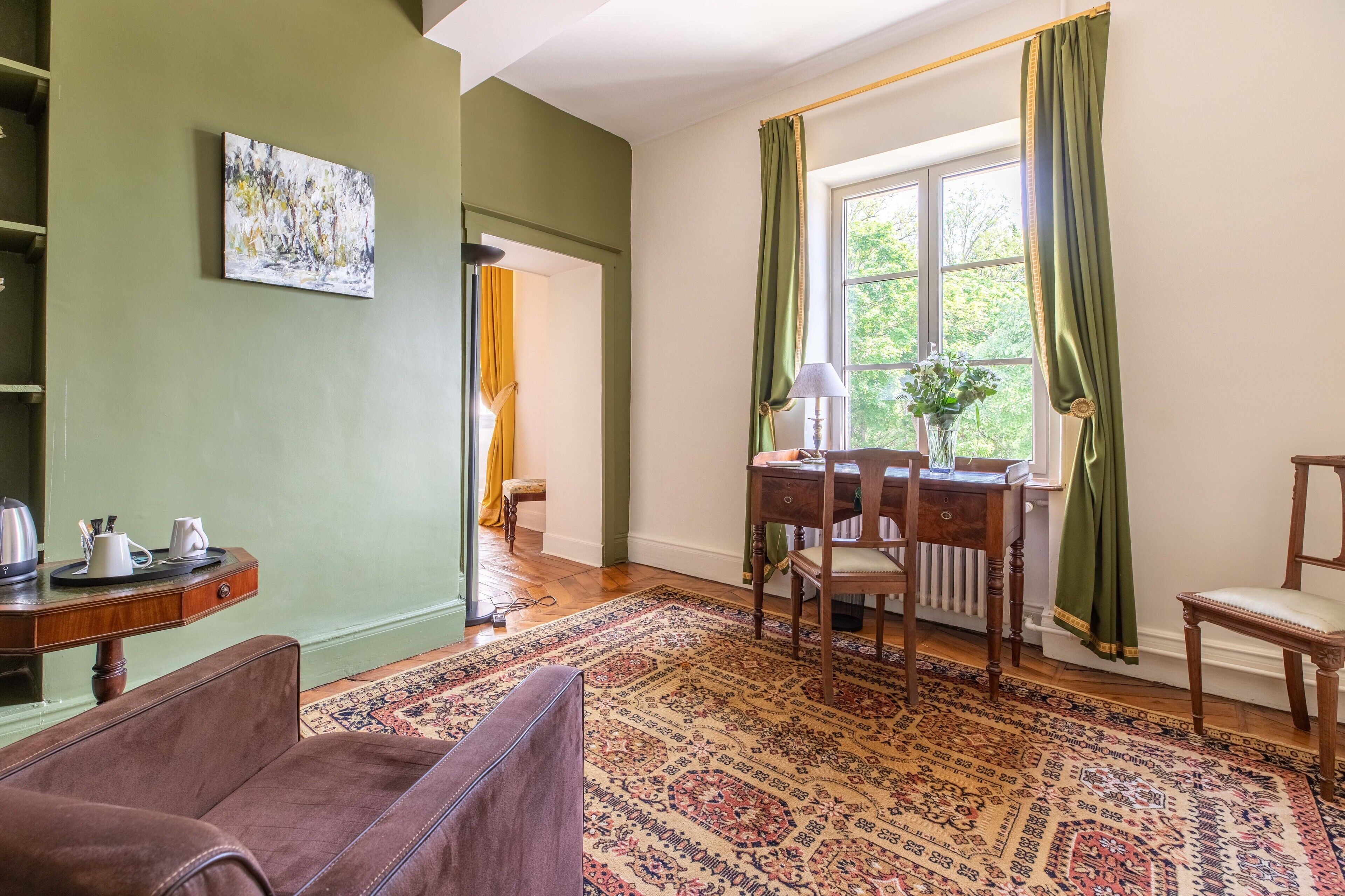 A room with a green wall, a wooden desk, a chair, a patterned rug, and a window with curtains.