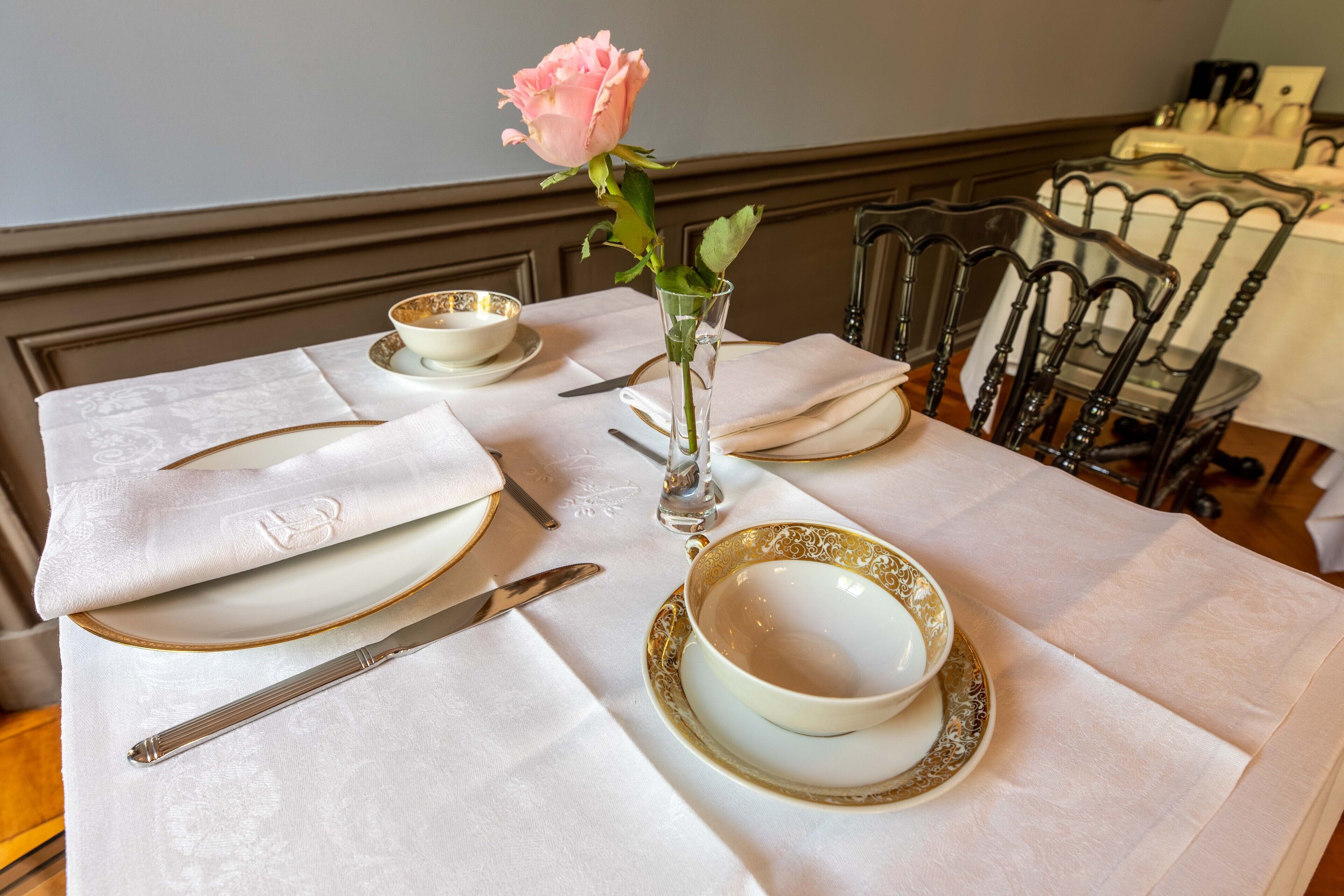 Una mesa de comedor dispuesta con platos blancos, cuencos con borde dorado y un florero de vidrio transparente con una rosa rosa.