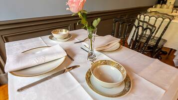 Una mesa de comedor dispuesta con platos blancos, cuencos con borde dorado y un florero de vidrio transparente con una rosa rosa.