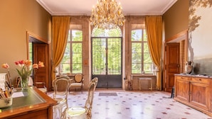 A spacious room with a large chandelier, a wooden desk, and a view of the outdoors through large windows. - Chambres D'hôtes Vichy : Château de Charmeil (Charmeil)