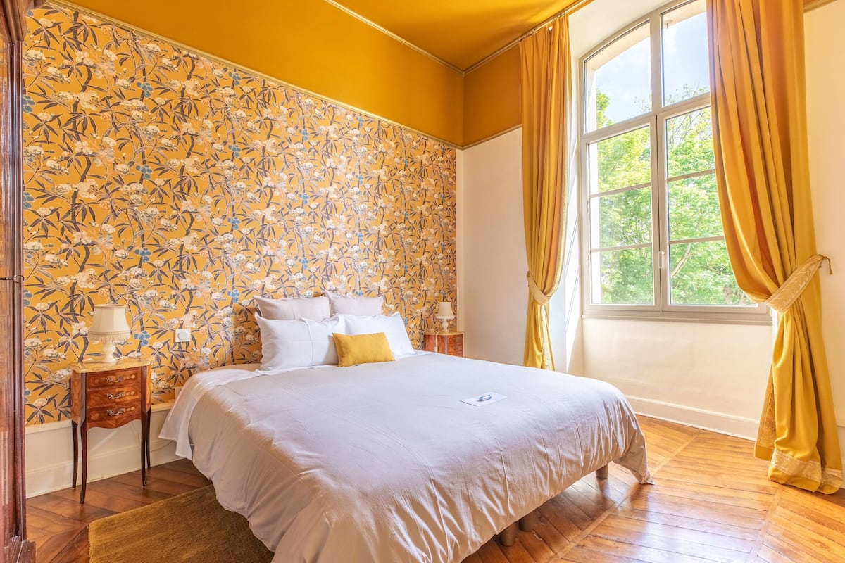 Ein Schlafzimmer mit floralem Wandpapier, einem großen Bett mit weißer Bettwäsche, einem Nachttisch mit Lampe und einem Fenster mit Vorhängen.