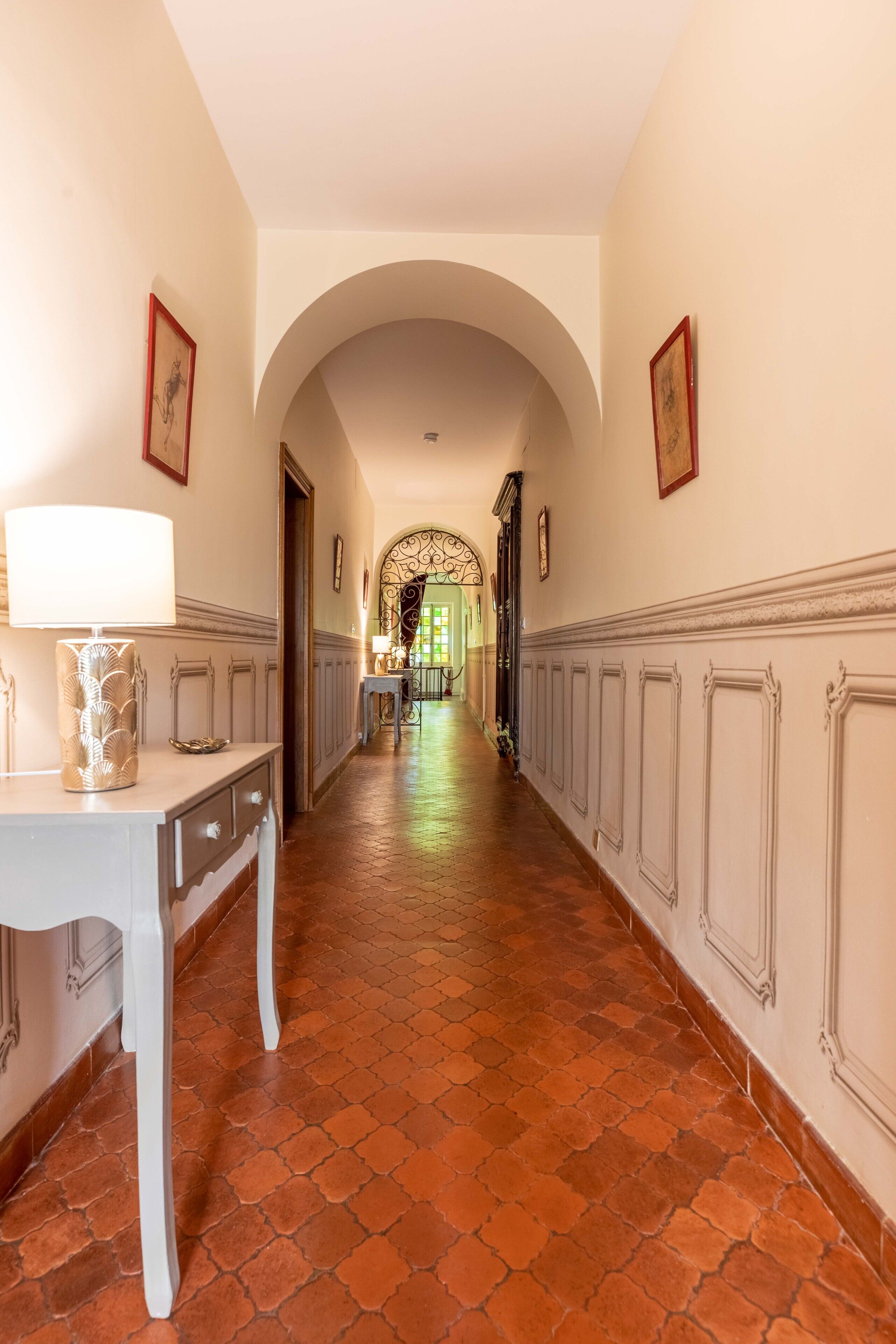 A hallway with a white table and lamp, tiled floor, and framed pictures on the walls.