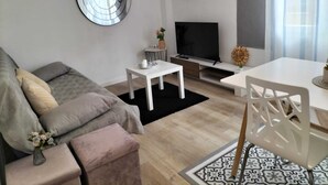 Una sala de estar moderna con un sofá gris, un televisor, una mesita y una zona de comedor con una mesa y sillas blancas.