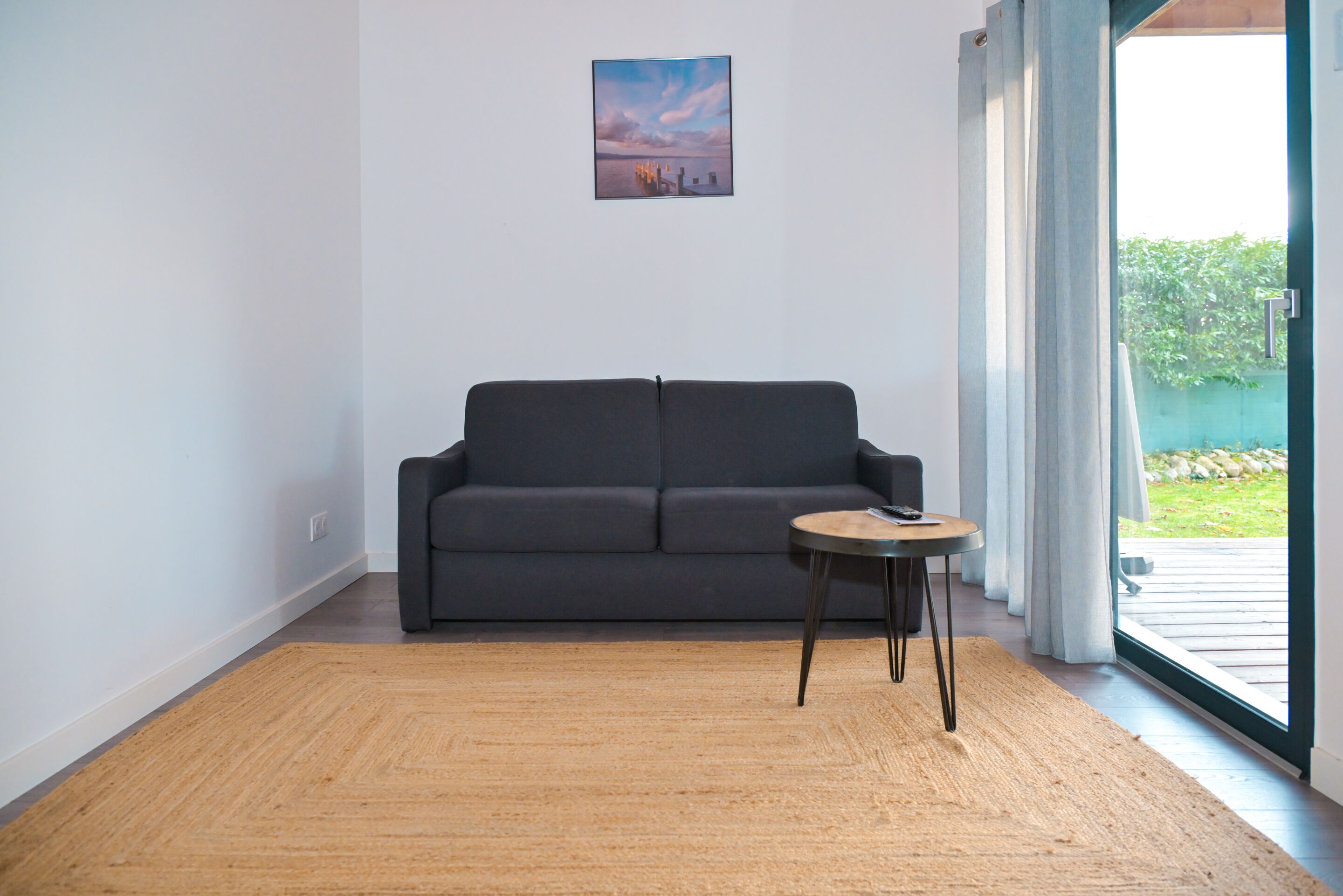 A living room with a dark grey sofa, a small round table, and a large window with curtains.