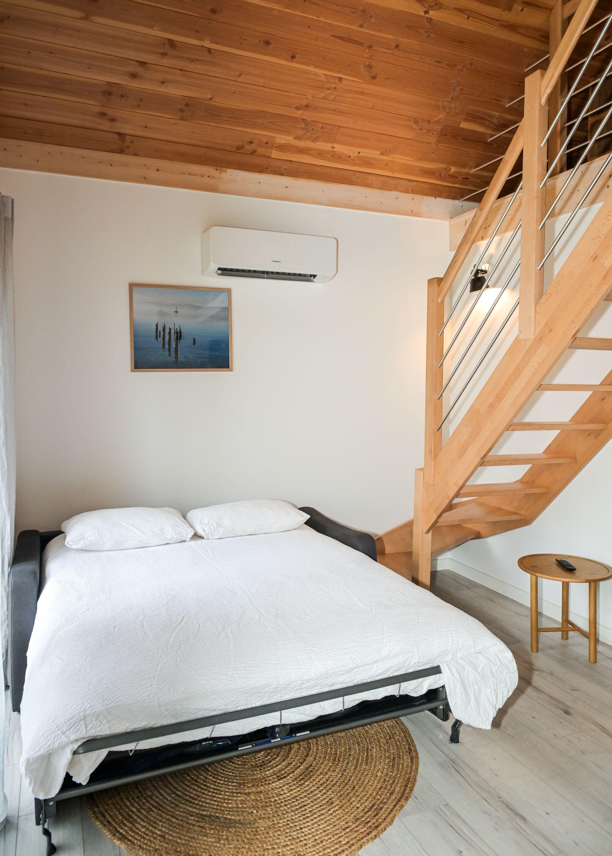 A bedroom with a bed, a painting, an air conditioning unit, a wooden staircase, and a small table.
