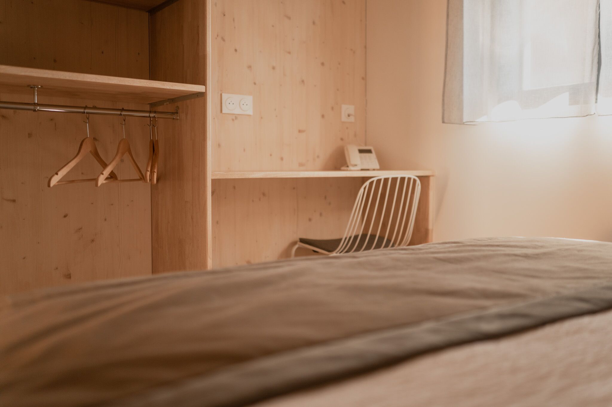 A bedroom with a bed, a wooden wardrobe, a chair, and a desk with a telephone.
